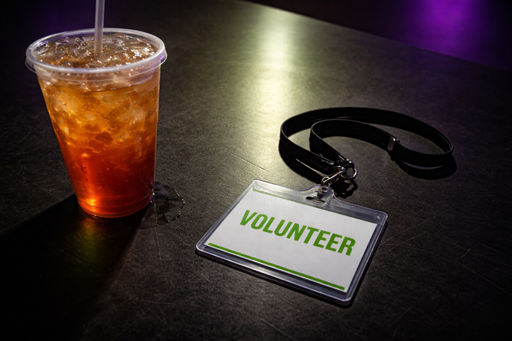 Extreme close-up, top-down shot of a conference table surface. A plastic iced tea cup sits next to a volunteer lanyard badge on a matte black table. The badge reads VOLUNTEER in green. 